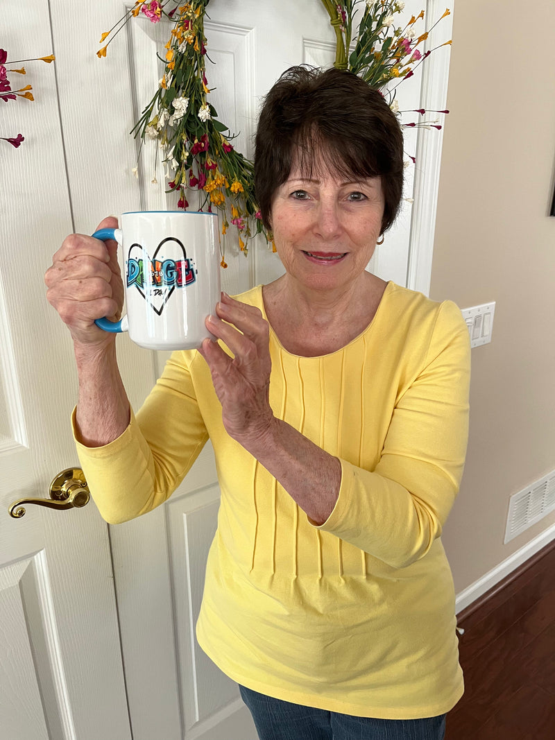 A woman holding a 15oz white ceramic mug with a blue handle and interior featuring a colorful Dance text and heart graphic