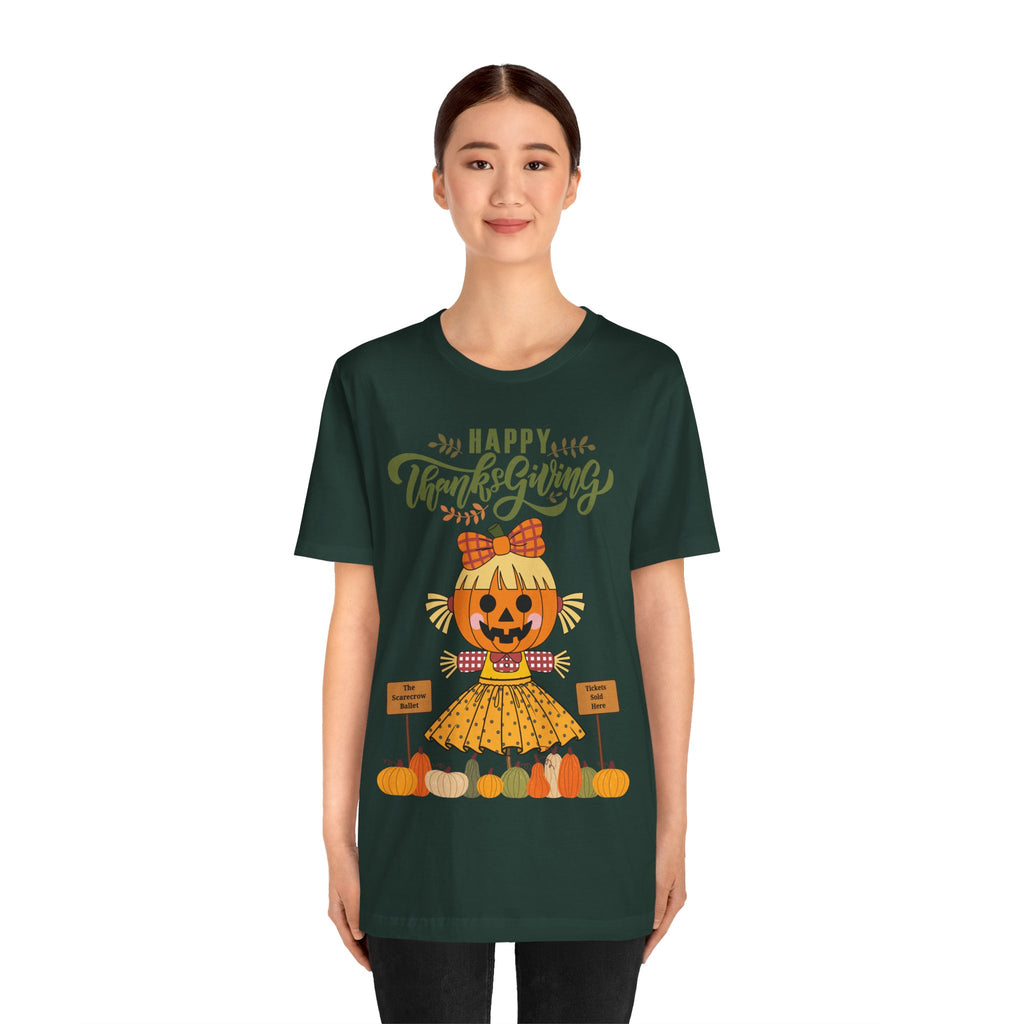 A woman wearing a forest green short-sleeve t-shirt with a Happy Thanksgiving graphic featuring a ballerina scarecrow design.