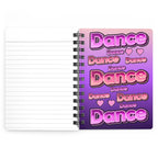 A spiral-bound lined notebook shown open, with a white lined page on the left and a cover design on the right featuring the word 'Dance' repeated in pink and purple bubble lettering over a pink-to-purple gradient background.