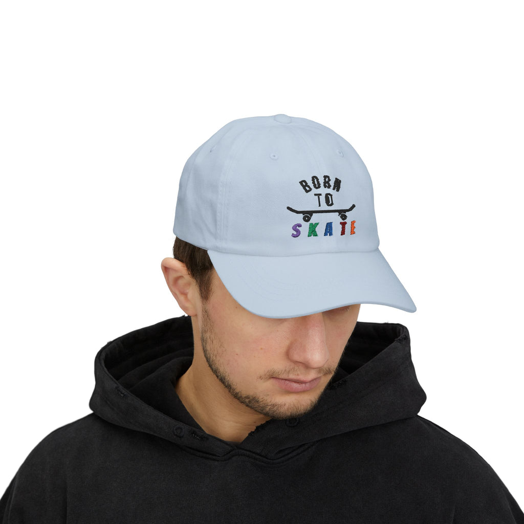 Man wearing a light blue baseball cap featuring a graphic on the front that reads BORN TO SKATE with a black skateboard silhouette and colorful lettering for SKATE.