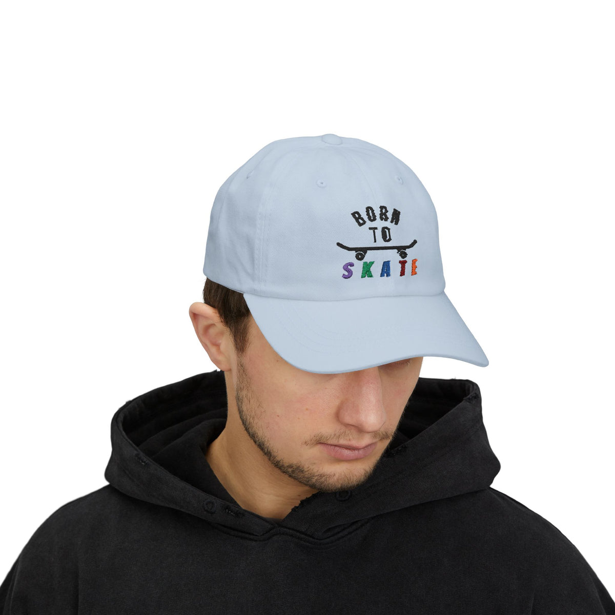 Man wearing a light blue baseball cap featuring a graphic on the front that reads BORN TO SKATE with a black skateboard silhouette and colorful lettering for SKATE.
