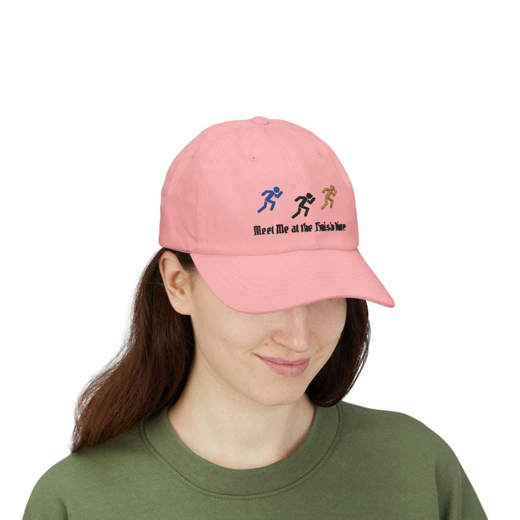 Woman wearing a pink baseball cap featuring embroidered running figures in blue, black, and tan above the text Meet Me at the Finish Line.