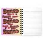 An open spiral-bound notebook with lined pages on the right and a decorative cover on the left featuring the word 'Dance' repeated in a retro gradient design.
