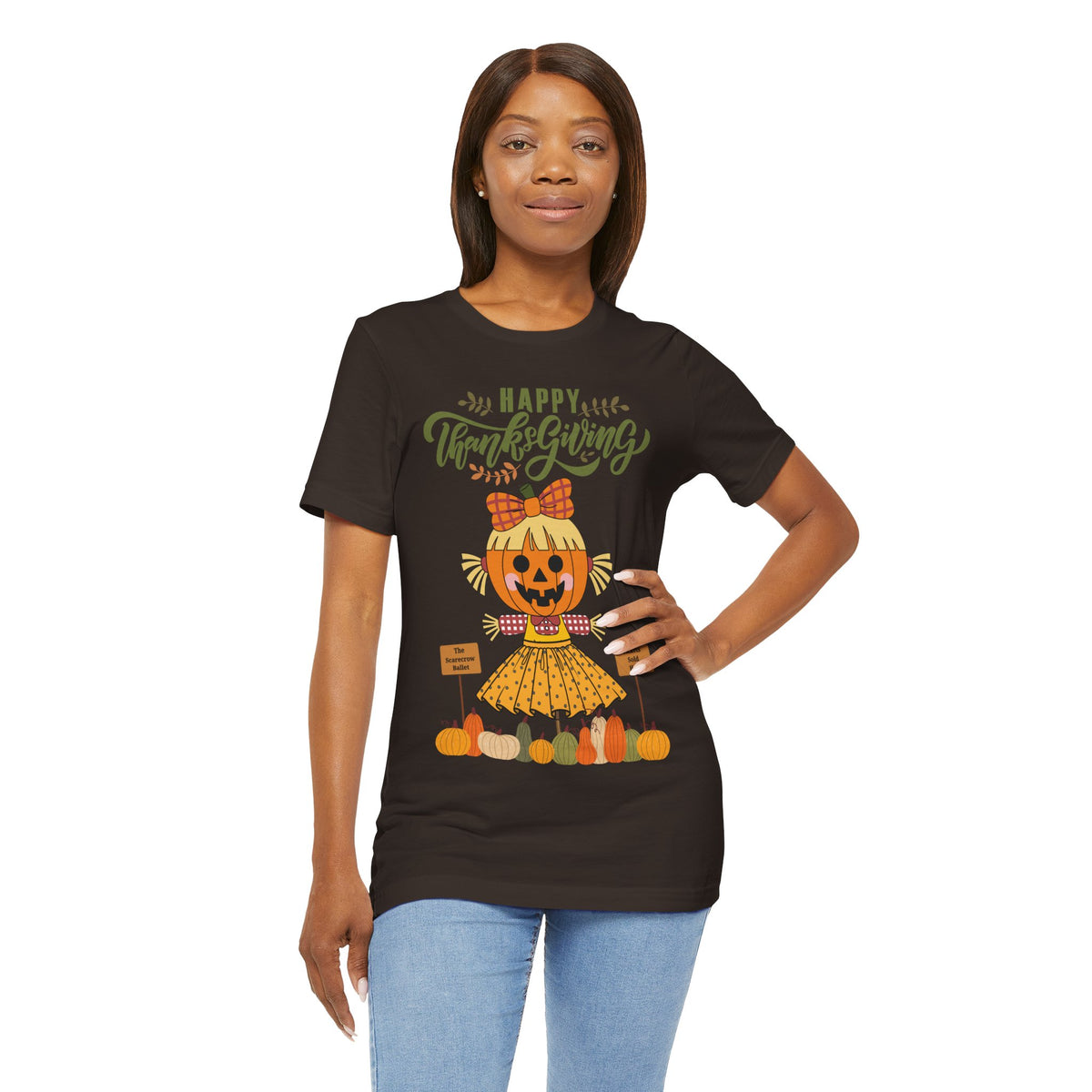 A woman wearing a brown short-sleeve t-shirt with a Happy Thanksgiving graphic featuring a ballerina scarecrow design.