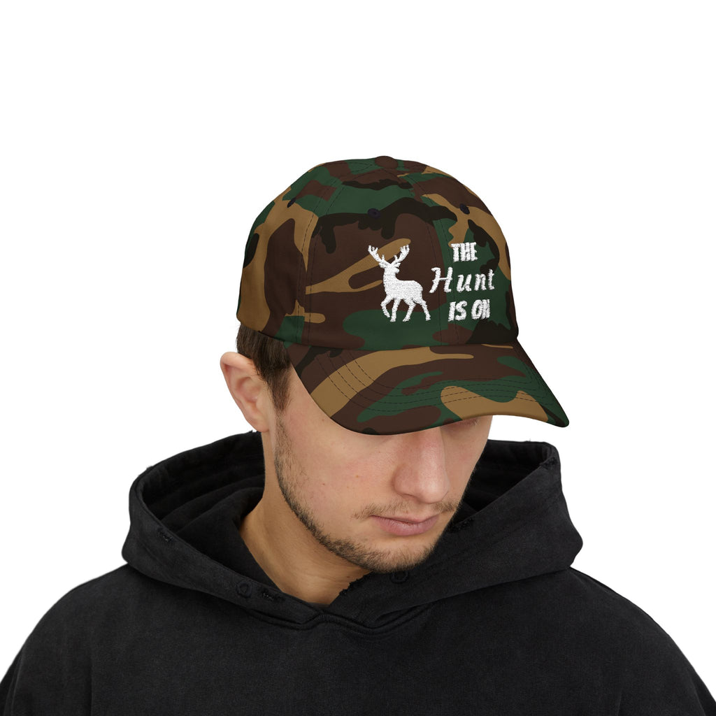 Man wearing a camouflage cap with an embroidered deer graphic and The Hunt Is On text.