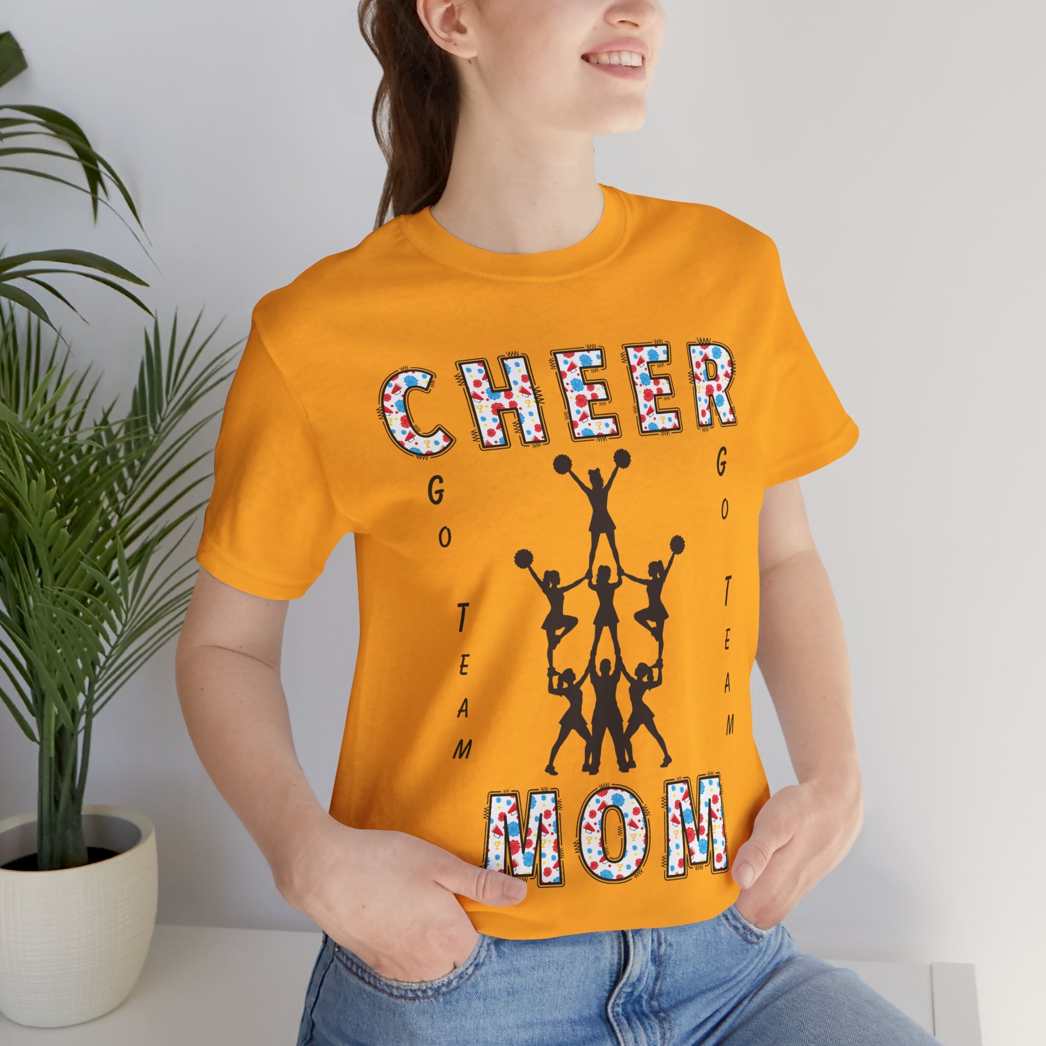 A woman wearing a gold graphic t-shirt with CHEER GO TEAM MOM text and graphic on the front.