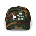 Camouflage cap with an embroidered deer graphic and The Hunt Is On text.