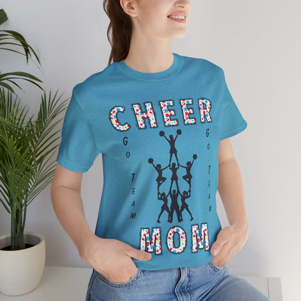 A woman wearing a heather aqua graphic t-shirt with CHEER GO TEAM MOM text and graphic on the front.