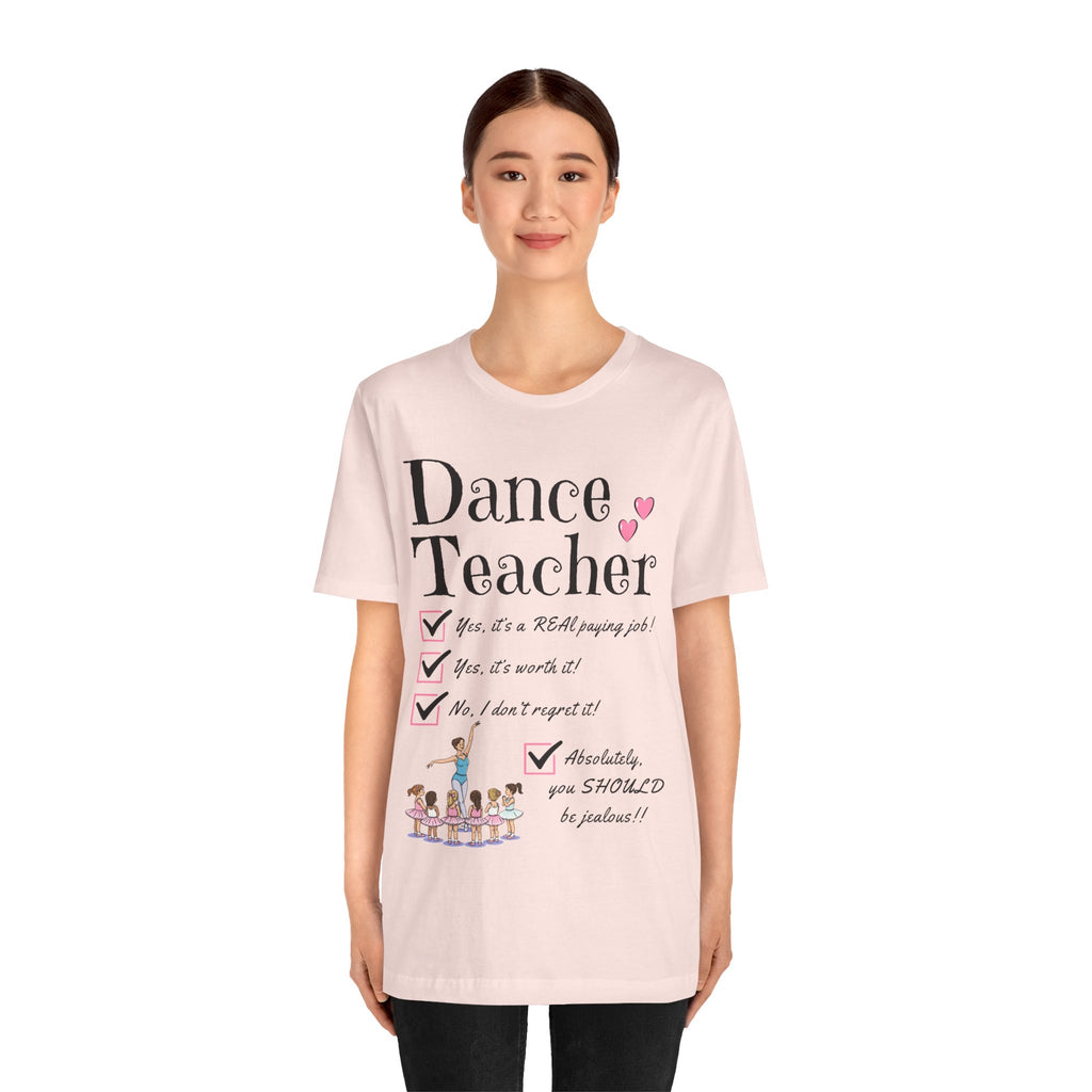A young woman wearing a soft pink short-sleeve crewneck t-shirt featuring a text that reads Dance Teacher and a ballet class graphic.