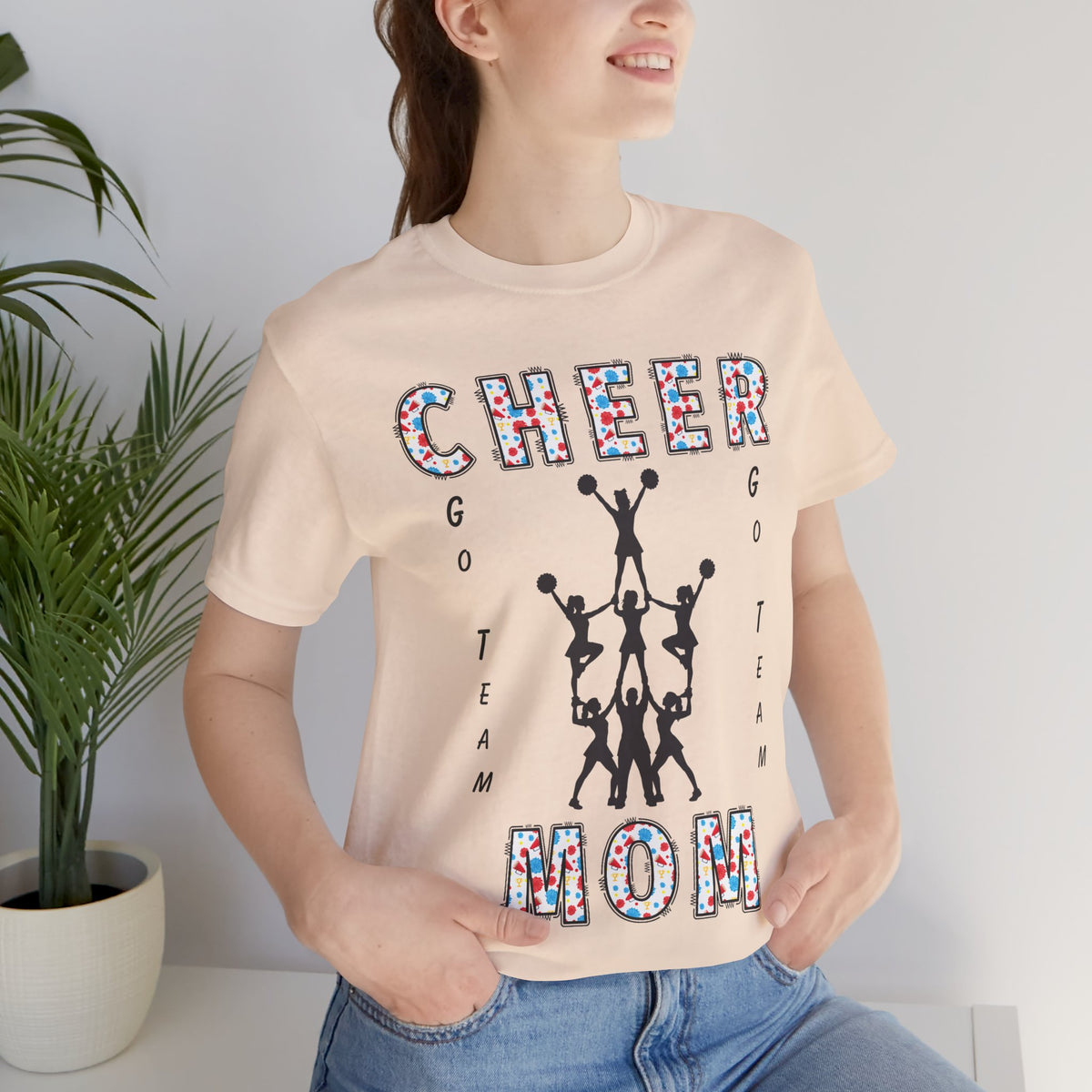 A woman wearing a soft cream graphic t-shirt with CHEER GO TEAM MOM text and graphic on the front.