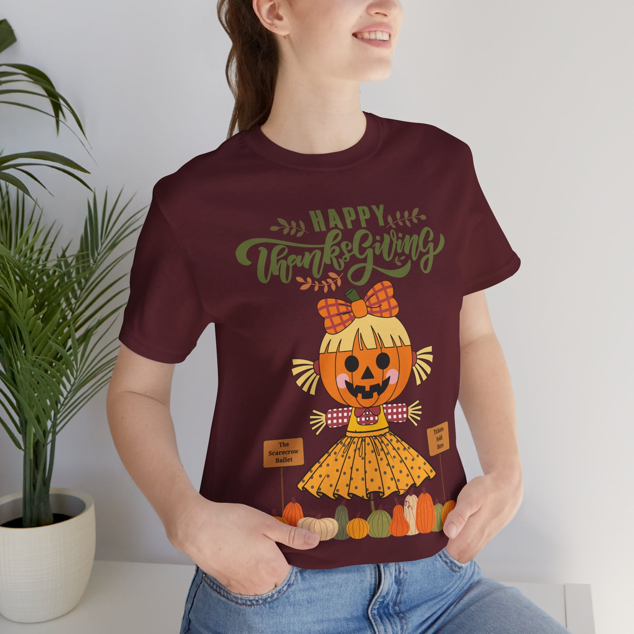 A woman wearing a maroon short-sleeve t-shirt with a Happy Thanksgiving graphic featuring a ballerina scarecrow design.