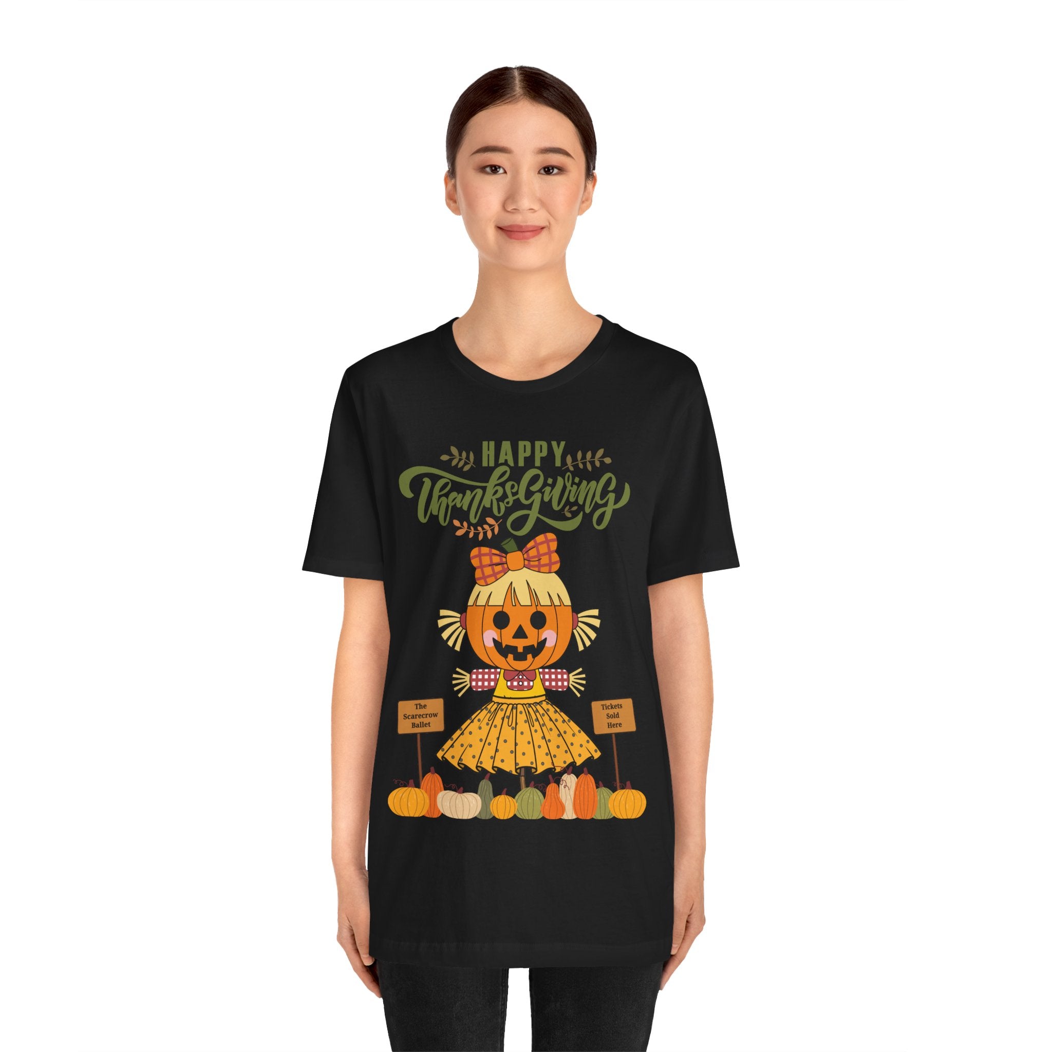 A woman wearing a black short-sleeve t-shirt with a Happy Thanksgiving graphic featuring a ballerina scarecrow design.