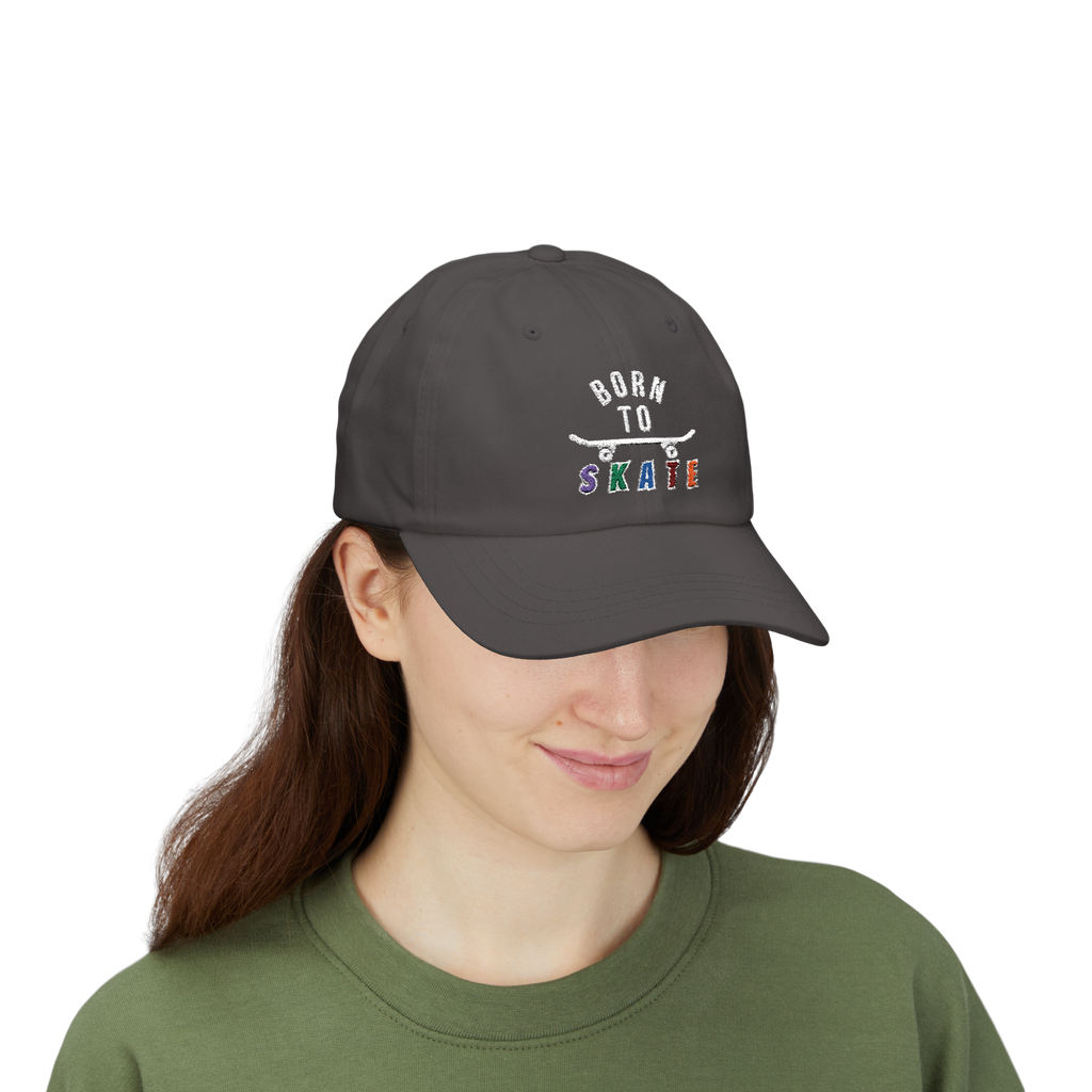 A woman wearing a dark grey baseball cap featuring a graphic on the front that reads BORN TO SKATE with a skateboard illustration. 